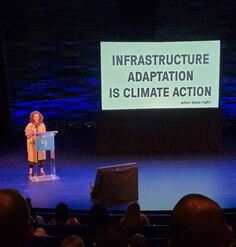 Anya Domlesky behind a podium on a stage with a large screen next turn her show a white slide with bold black words "INFRASTRUCTURE ADAPTATION IS CLIMATE ACTION when done right"