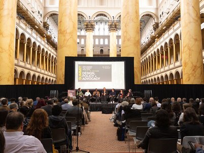 The stage, screen, and panelists in front of a seated audience amidst the columns and arches of the National Building Museum's Great Hall